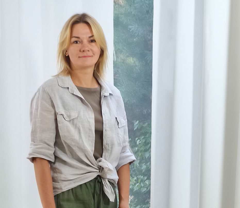 Kobieta o blond włosach stoi w pomieszczeniu przed białymi zasłonami i oknem z zielenią na zewnątrz. Ma na sobie jasną koszulę przewiązaną w pasie, szary top i zielone spodnie, i patrzy w kamerę z lekkim uśmiechem.