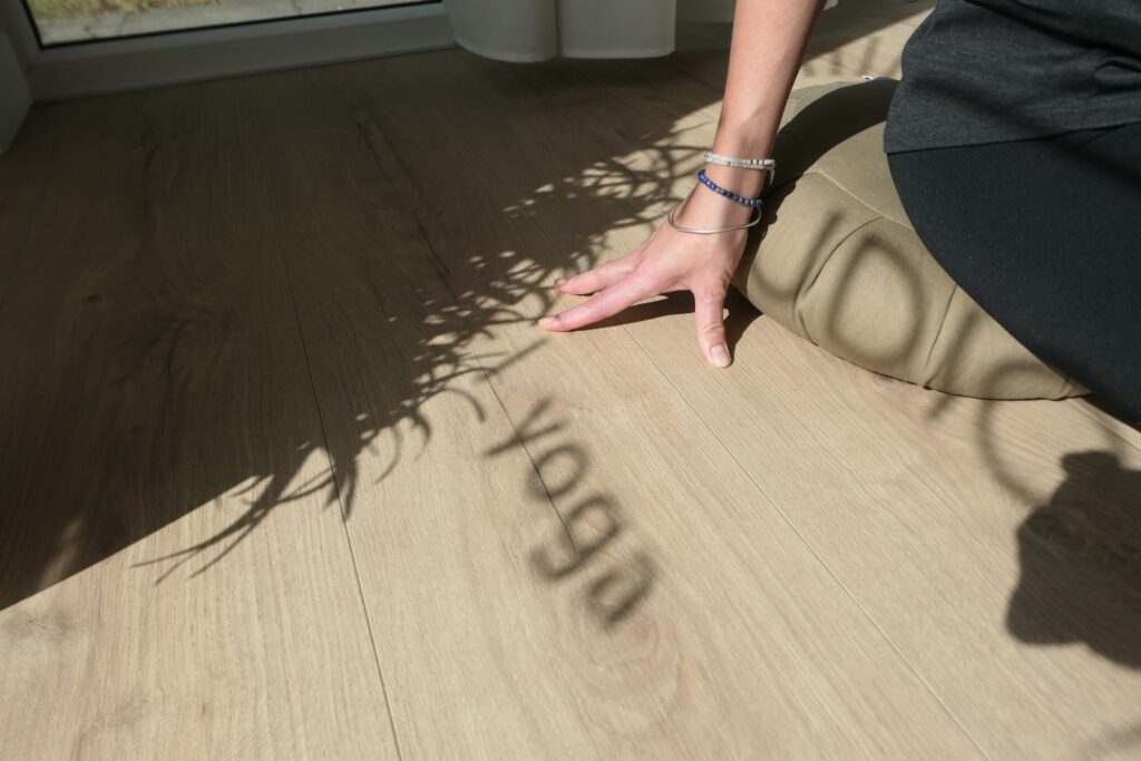 Osoba siedzi na poduszce do jogi na drewnianej podłodze, a światło słoneczne rzuca cień słowa "joga" i rośliny na podłogę; widoczna jest tylko jej ręka i część ciała.