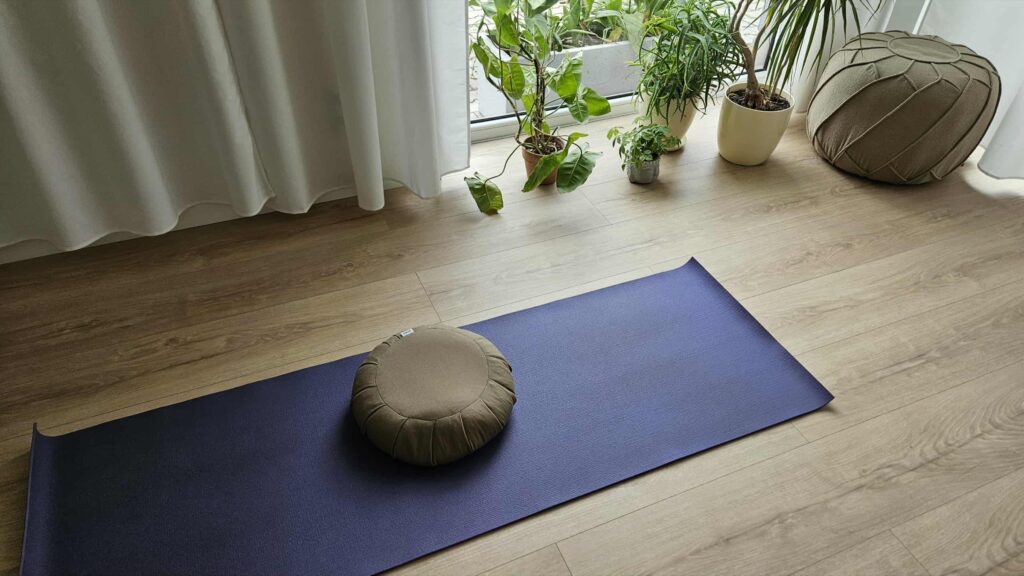 Niebieska mata do jogi z okrągłą poduszką do medytacji jest umieszczona na drewnianej podłodze w pobliżu okna z kilkoma roślinami doniczkowymi i pufą. Białe zasłony częściowo zakrywają okno.