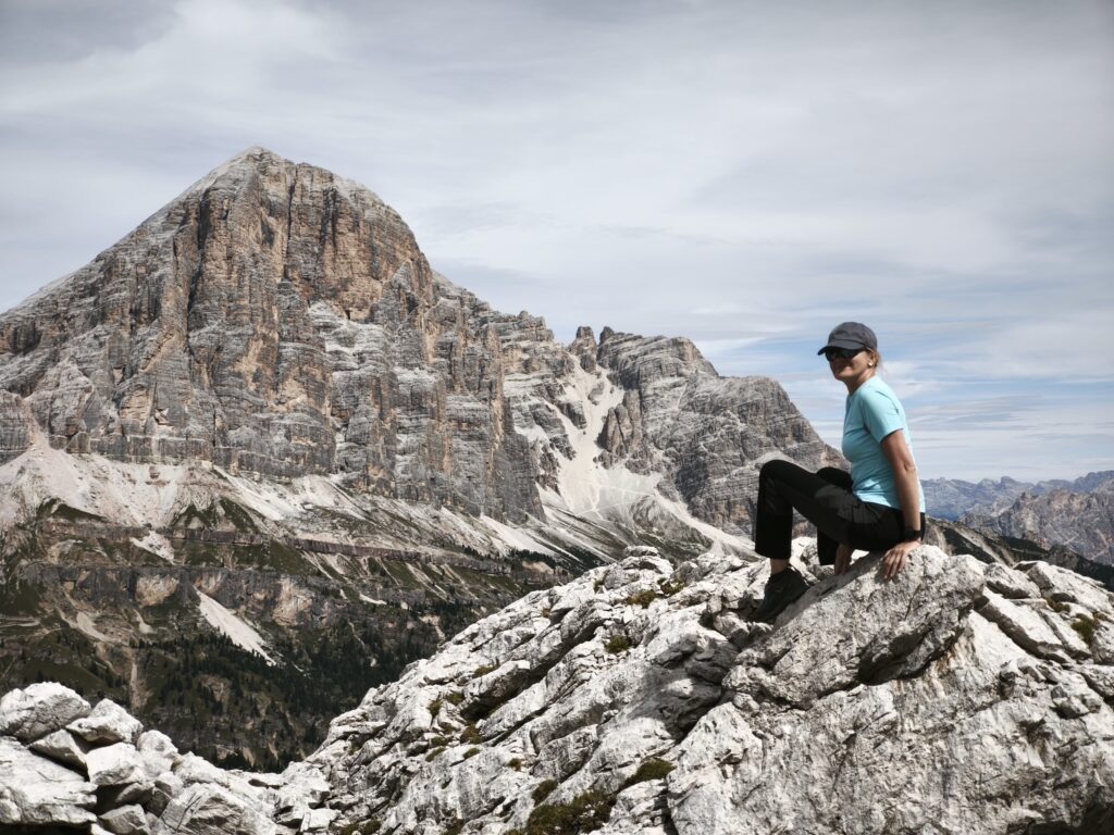 Osoba w niebieskiej koszuli i czapce siedzi na skalistym terenie, z widokiem na dramatyczny górski krajobraz z surowymi szczytami i zachmurzonym niebem w tle.