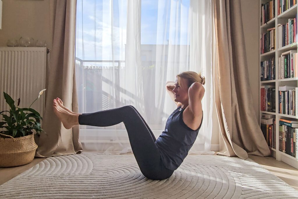 Kobieta w stroju treningowym wykonuje ćwiczenia brzucha na jasnym dywanie w oświetlonym słońcem pokoju, z przezroczystymi zasłonami, rośliną doniczkową i półką z książkami w tle - idealne miejsce na codzienną sesję jogi online.