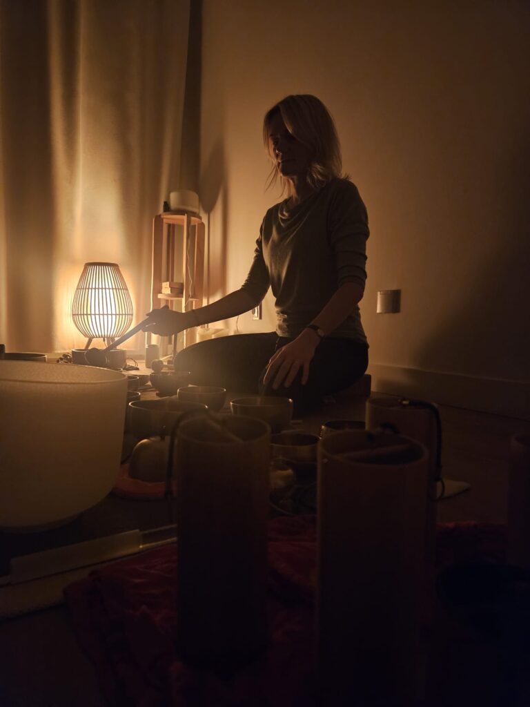 Kobieta siedzi ze skrzyżowanymi nogami na podłodze w słabo oświetlonym pokoju, oświetlonym ciepłą lampą, otoczona śpiewającymi misami i instrumentami do uzdrawiania dźwiękiem. Wygląda na spokojną i skupioną podczas medytacji lub sesji terapii dźwiękiem.