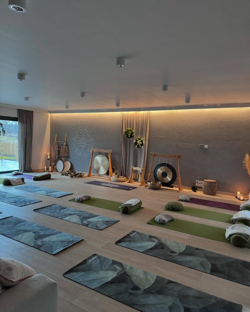 Spokojne studio jogi z matami, podpórkami i poduszkami starannie ułożonymi na drewnianej podłodze. Gongi, świece i naturalne dekoracje ustawione są z przodu, a delikatne oświetlenie tworzy spokojną, relaksującą atmosferę.