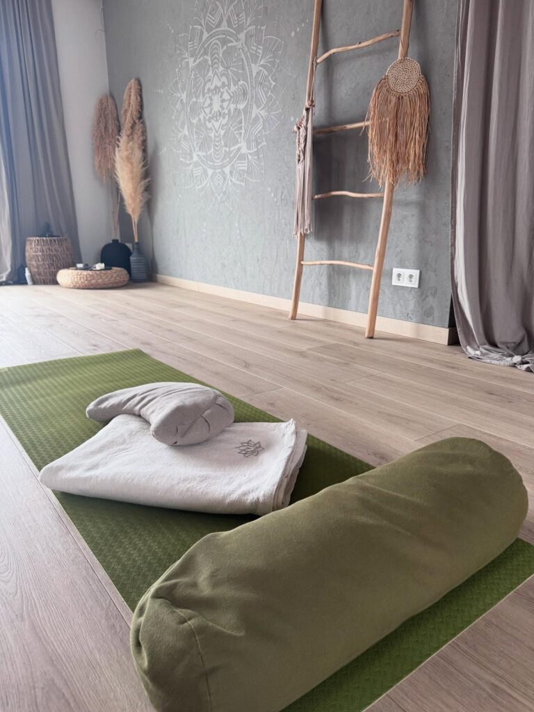 Zielona mata do jogi z białym ręcznikiem, podpórką i złożonym kocem leży na jasnej drewnianej podłodze w spokojnym studio. Dekoracyjna drabina, grafika ścienna i trawa pampasowa dodają spokojnego, minimalistycznego wystroju.