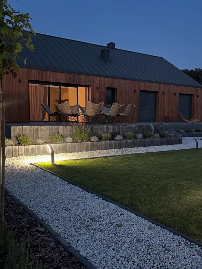 Nowoczesny dom z drewnianym sidingiem i dużymi oknami jest oświetlony od wewnątrz o zmierzchu. Na patio znajdują się plecione krzesła i stół z widokiem na ogród z białym żwirem, zielonym trawnikiem i świecącym oświetleniem ścieżki.