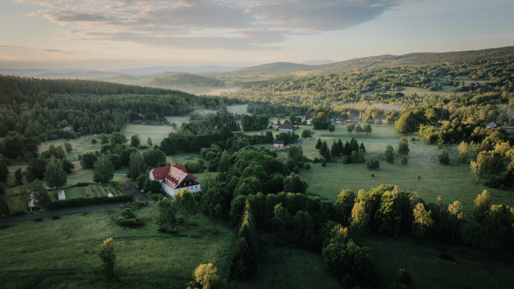 Widok z lotu ptaka na bujną, zieloną okolicę z rozrzuconymi drzewami, polami i budynkami, w tym biały dom z czerwonym dachem, otoczony lasami i falistymi wzgórzami pod częściowo zachmurzonym niebem.