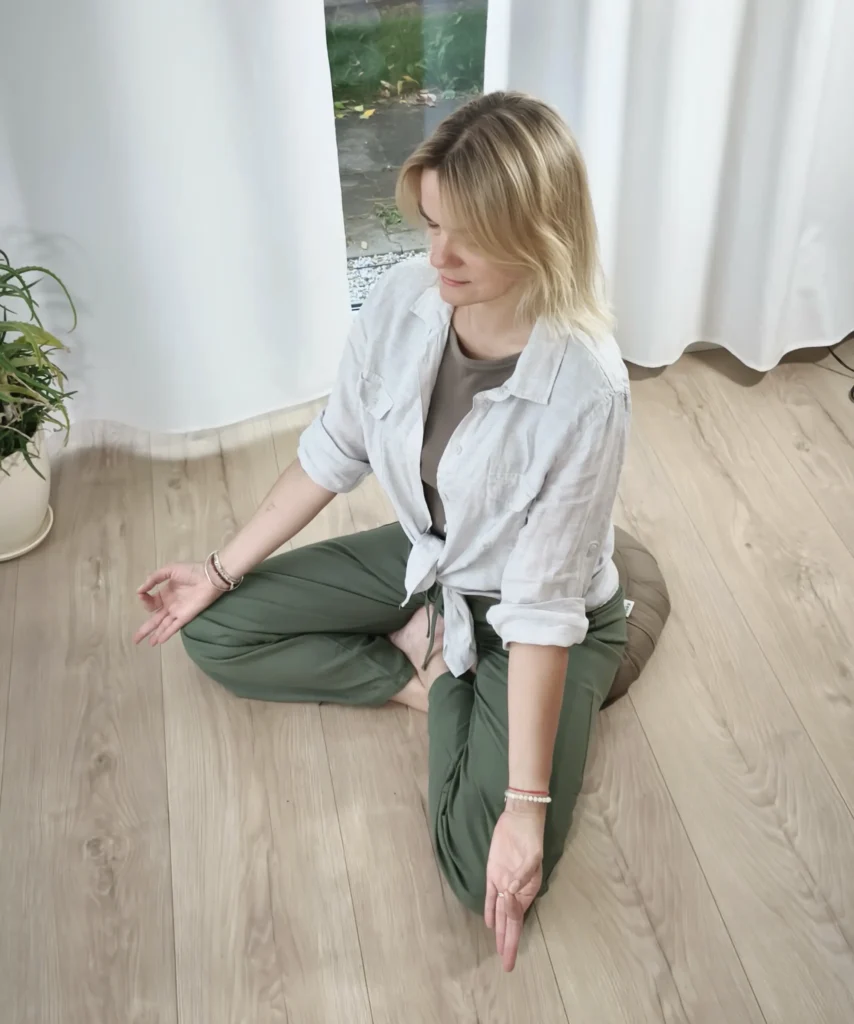 Kobieta o blond włosach siedzi ze skrzyżowanymi nogami na poduszce w jasnym pokoju, medytując w ramach warsztatu uważności, a jej ręce spoczywają na kolanach, dłońmi do góry, obok rośliny doniczkowej na drewnianej podłodze.