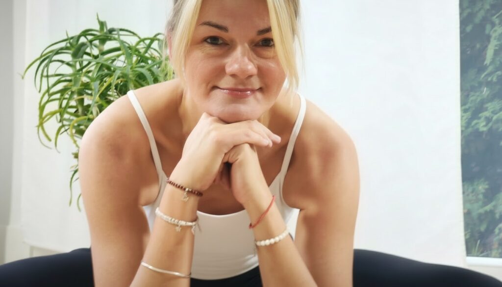 Kobieta w białym podkoszulku uśmiecha się i opiera podbródek na dłoniach, pochylając się do przodu w pomieszczeniu. W tle widoczna jest zielona roślina.