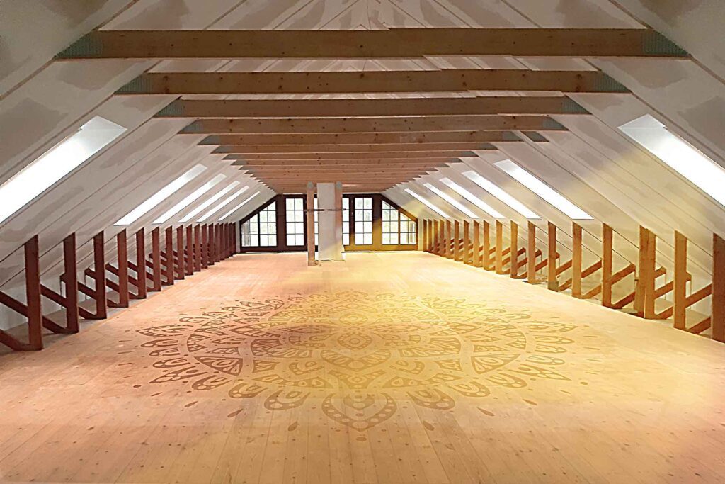 Przestronne, wypełnione światłem poddasze z pochyłymi białymi sufitami, drewnianymi belkami i szczegółowym okrągłym wzorem mandali na drewnianej podłodze. Duże okna znajdują się wzdłuż przeciwległej ściany, dzięki czemu naturalne światło wypełnia pomieszczenie.