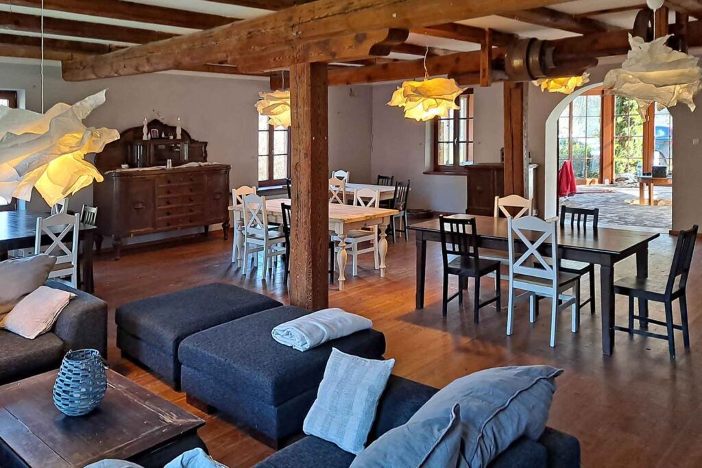 Przestronny salon i jadalnia z odsłoniętymi drewnianymi belkami, ciemnymi sofami z poduszkami, drewnianymi stołami i krzesłami, wiszącymi lampami wiszącymi z pomarszczonymi białymi kloszami oraz światłem słonecznym wpadającym przez duże okna.