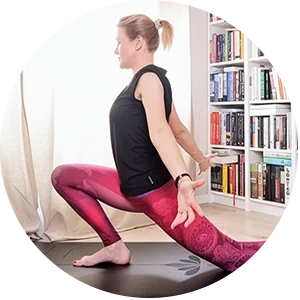 Kobieta ćwiczy jogę w pomieszczeniu, wykonując pozycję do wykroku na macie. Ma na sobie czarny top i różowe legginsy, a w tle półkę wypełnioną książkami i światło słoneczne wpadające przez okno.