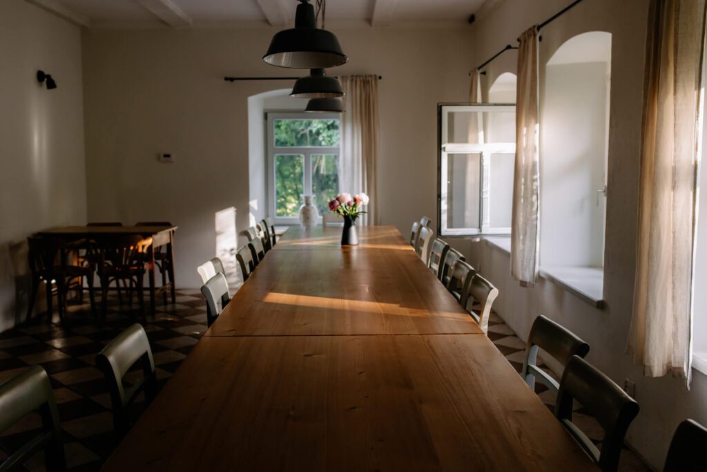 Długi drewniany stół z krzesłami w oświetlonym słońcem pokoju, wazon z kwiatami pośrodku. Duże okna z kremowymi zasłonami wpuszczają naturalne światło. W tle widoczne są dwa mniejsze stoły i krzesła.
