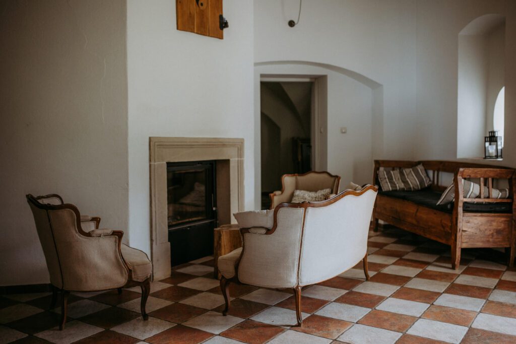 Przytulny salon z zabytkowymi fotelami i małą sofą wokół kominka, na podłodze wyłożonej brązowymi i białymi kafelkami w kratkę, z łukowatymi drzwiami i ścianami w neutralnych kolorach.