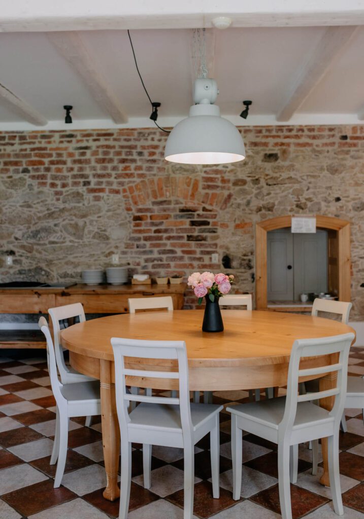 Okrągły drewniany stół z sześcioma białymi krzesłami, czarny wazon z różowymi kwiatami pośrodku, znajduje się w rustykalnym pokoju z ceglanymi i kamiennymi ścianami, podłogą wyłożoną kafelkami w kratkę i dużą białą lampą wiszącą nad głową.