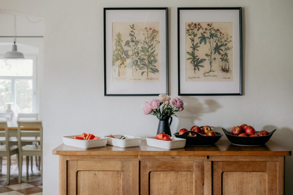 Drewniany kredens z miskami jabłek, tacami z krojonymi owocami i wazonem z różowymi kwiatami, pod dwoma oprawionymi wydrukami botanicznymi na białej ścianie. W tle widoczny jest stół i krzesła.