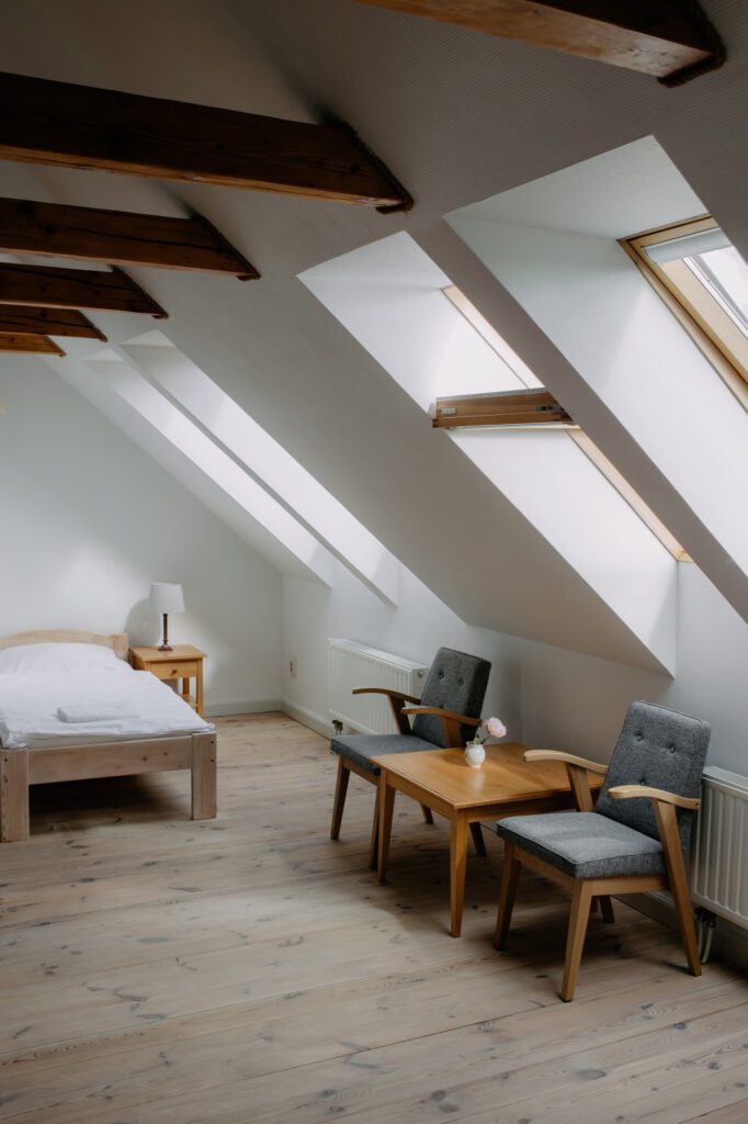 Jasna sypialnia na poddaszu z drewnianymi belkami, pojedynczym łóżkiem, małym stolikiem z dwoma szarymi krzesłami, lampą i dużymi oknami wpuszczającymi naturalne światło na jasne drewniane podłogi.