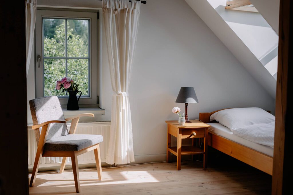 Przytulna sypialnia z drewnianym łóżkiem, szarym krzesłem, szafką nocną z lampką i kwiatami oraz oknem z białymi zasłonami wpuszczającymi naturalne światło. Na parapecie stoi czarny wazon z różowymi kwiatami.