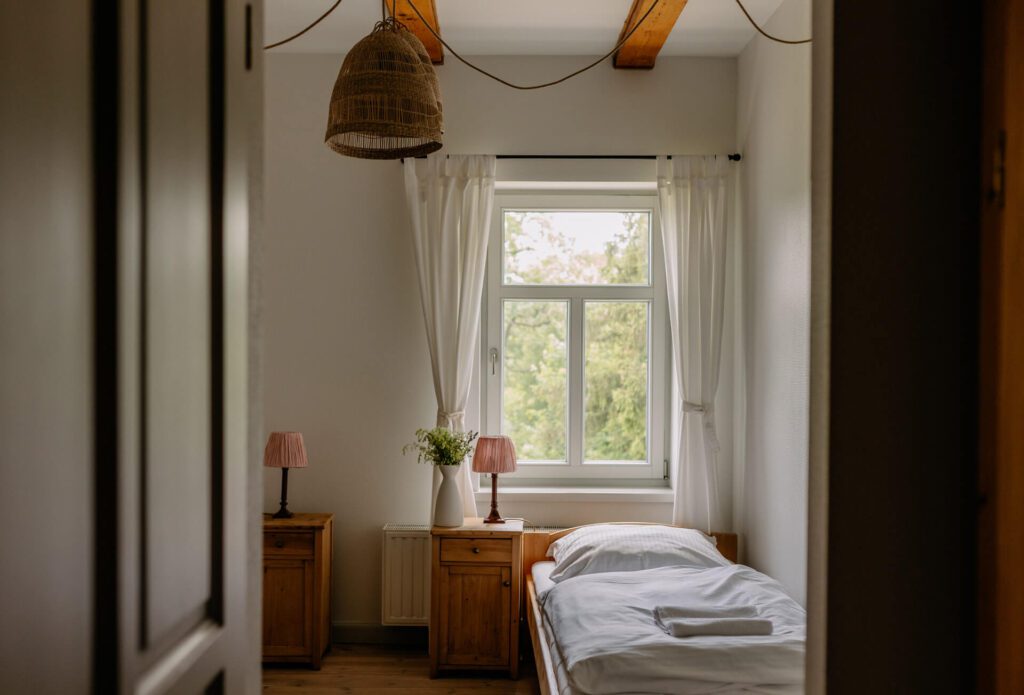 Przytulna sypialnia z pojedynczym łóżkiem, białą pościelą, dwiema drewnianymi szafkami nocnymi z lampkami, oknem z białymi zasłonami i wiszącym wiklinowym abażurem. Światło słoneczne wpada do środka, tworząc spokojną, zachęcającą atmosferę.