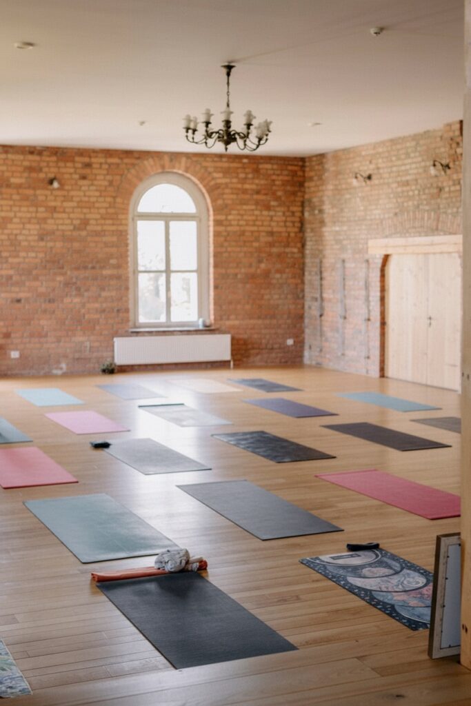 Przestronny pokój z drewnianymi podłogami i odsłoniętymi ceglanymi ścianami jest przygotowany na zajęcia jogi. Kilka kolorowych mat do jogi ułożonych jest starannie na podłodze, a obok nich znajdują się rekwizyty do jogi. Duże okno wpuszcza naturalne światło.