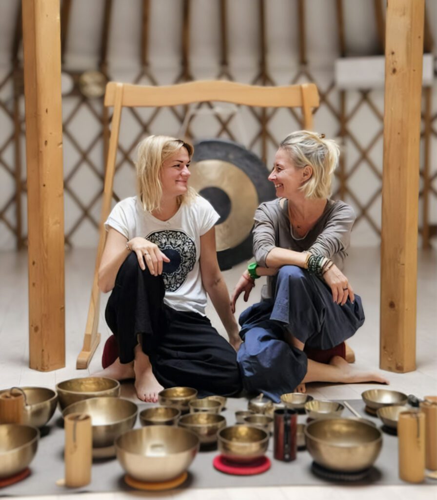 Dwie kobiety siedzą ze skrzyżowanymi nogami na podłodze, uśmiechając się do siebie, otoczone różnymi śpiewającymi misami i instrumentami. Za nimi wisi duży gong, a drewniane belki otaczają spokojne wnętrze.