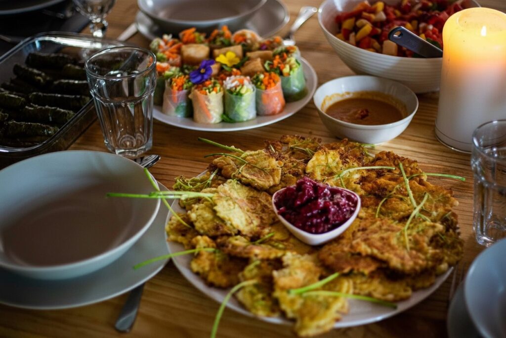 Drewniany stół nakryty do posiłku z talerzami z bułeczkami warzywnymi, liśćmi winogron, latkes z sosem buraczanym, miską sałatki owocowej, szklankami i zapaloną świecą.