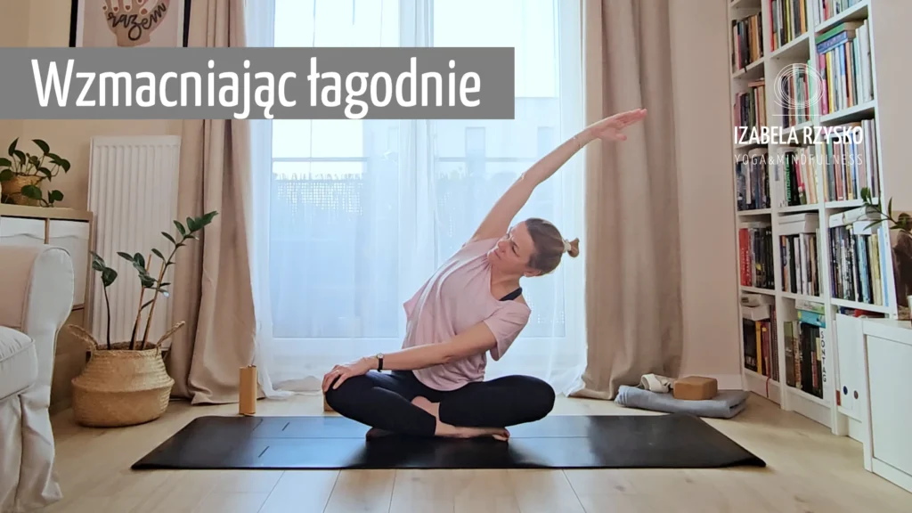 Kobieta siedzi ze skrzyżowanymi nogami na macie do jogi w pomieszczeniu, wyciągając prawą rękę w lewo. Tekst "Wzmacniając łagodnie" pojawia się w lewym górnym rogu, z jasnymi zasłonami i półkami z książkami w tle.