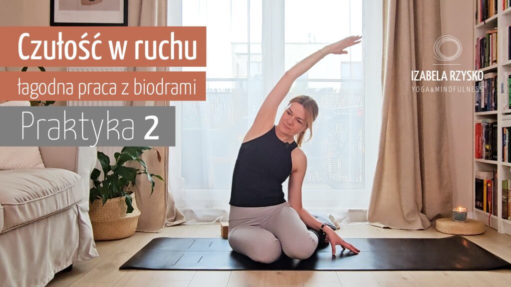 Kobieta siedzi na macie do jogi w pomieszczeniu, wyciągając lewą rękę nad głowę. Tekst w języku polskim brzmi: Czułość w ruchu, łagodna praca z biodrami, Praktyka 2. Delikatnie oświetlony pokój z roślinami i wystrojem tworzy scenę dla praktyki jogi online.