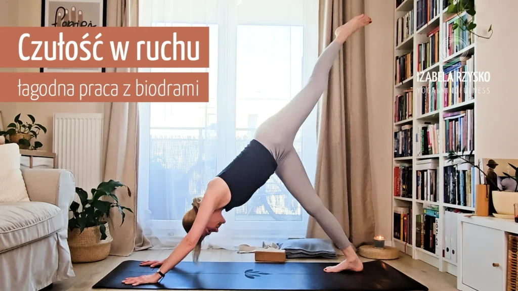 Iza ćwiczy joga online w pomieszczeniu na czarnej macie, balansując na rękach i jednej stopie z drugą nogą uniesioną wysoko. Znajduje się w jasnym pokoju z półką na książki, roślinami, miękkim oświetleniem, a na obrazie pojawia się polski tekst.