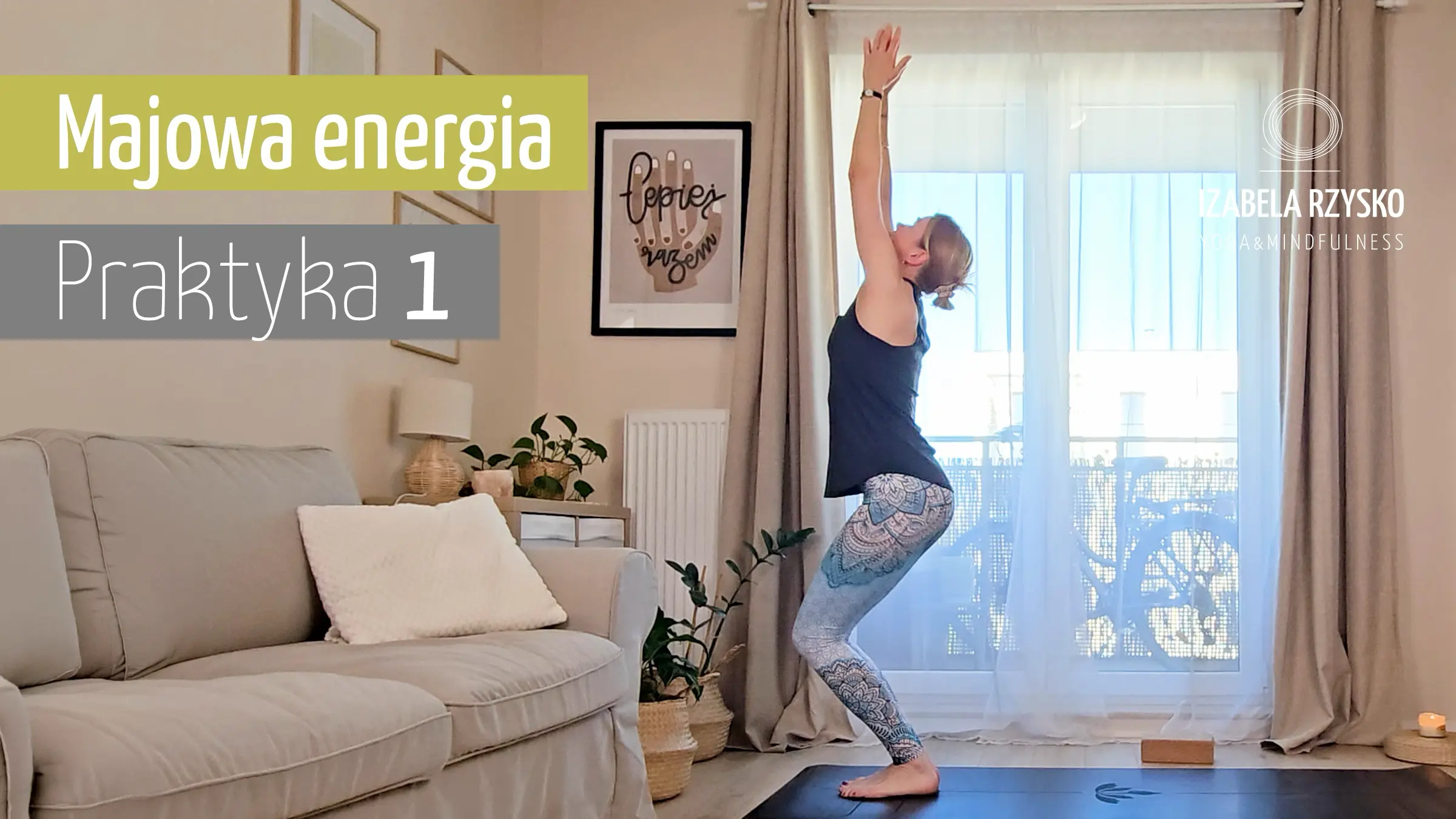 Kobieta ćwiczy jogę w pomieszczeniu, stojąc na macie w Utkatasana (pozycja krzesła) z podniesionymi rękami. Jasny pokój ma duże okna, sofę, rośliny i wystrój. Tekst brzmi: Majowa energia Praktyka 1, Izabela Rzysko Mindfulness, joga online.
