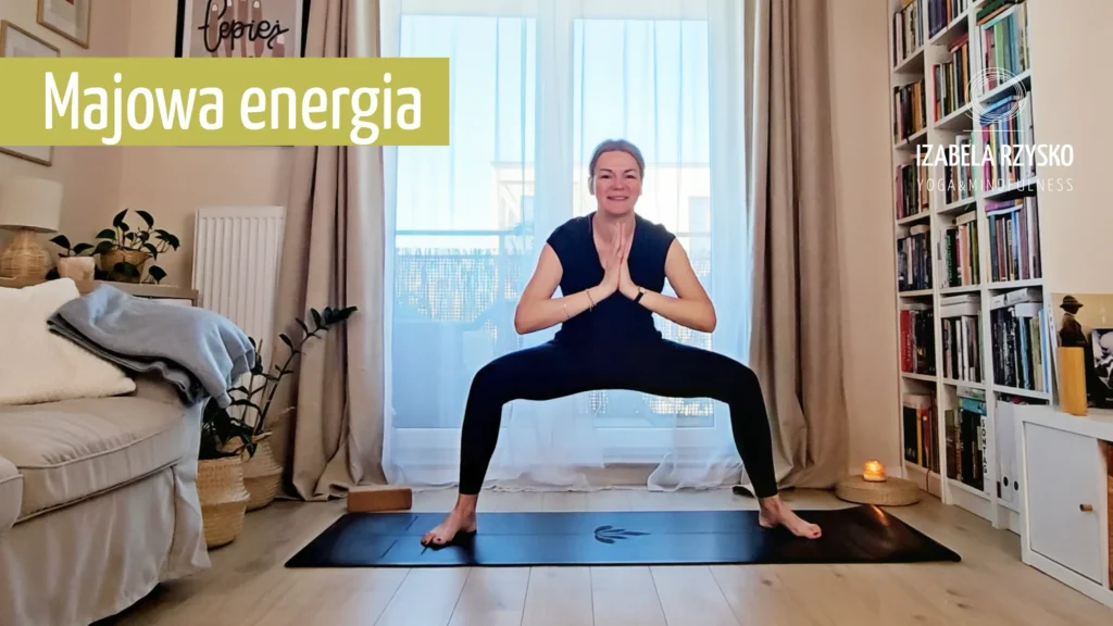 Iza Rzysko ćwiczy jogę online w dobrze oświetlonym salonie, stojąc w szerokim przysiadzie z rękami w modlitwie. Tekst na obrazie brzmi "Majowa energia" i "Izabela Ryszko Fitness". Widoczne są rekwizyty do jogi i półki z książkami.