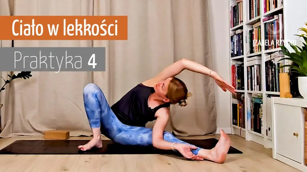 Iza Rzysko ćwiczy jogę online w salonie, siedzi na macie i rozciąga się w bok nad wyprostowaną nogą. W tle widać regał z książkami.